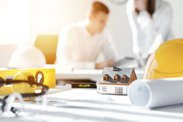 Fototapeta premium Selective focus. Close up shot of scale model house on table with yellow helmets and engineering tools, with two architects in background discussing work issues using electronic tablet during meeting