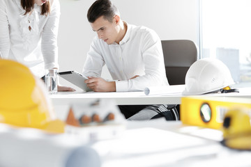 Two engineers or designers working on construction project, analyzing blueprint on digital tablet, discussing plans in office with scale model house, helmets and architectural tools in foreground