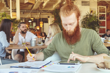 Bearded student working on course paper, making research, analyzing statistics on tablet, writing down in notebook using pencil. Redhead entrepreneur sitting at cafe with touch pad during lunch break
