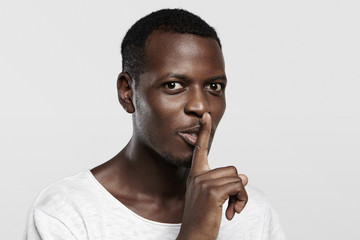 Fototapeta na wymiar Body language. Young African man in white t-shirt holding finger on his lips, asking to keep silence about confidential information, saying 'shh', looking at camera with serious face expression