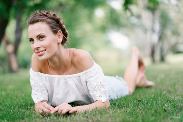 Naklejka na ściany i meble Young beautiful sexy girl lying on the grass in the park and rest. Beautiful healthy smile. Fancy Hairstyle. Happy girl