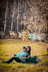 Young beautiful couple kissing and hugging near trees with blossom in summer park near stump on the grass. Woman in green dress and wreath. Man in blue shirt and gray waistcoat