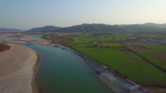 Backwards flying aerial shot over blue river in rural area of India.  Rocky riverbed, and lush green fields with clear skies.