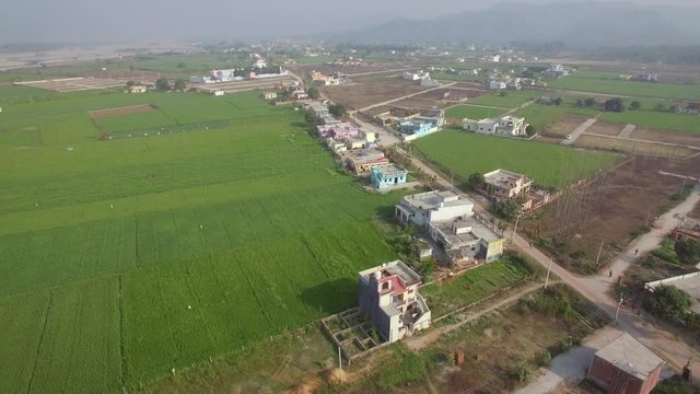 Aerial shot flying over agricultural fields in a rural Indian village. Hazy/dusty sky with hills in the distance. Lush green fields with scattered village houses along a small road. Turning shot.