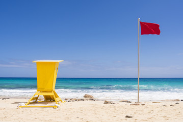 Yellow life guard station with red flag on sandy white beach.