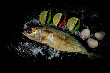 Grilled mackerel on a black background with lemon, salt, pepper, onion, and scallions.