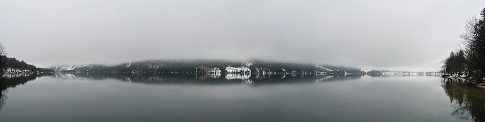Panoramic image of snowy, winter lake landscape, with pine trees reflecting on the water, on a cloudy day with dense fog. Nature with no people. Horizontal panorama. Bohinj lake, Slovenia. - 121695670