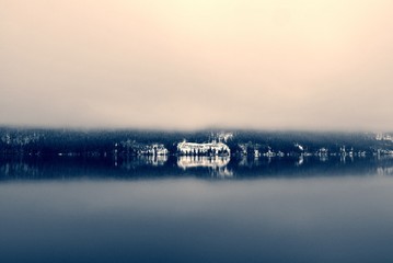 Snowy winter landscape on the lake in black and white, with trees reflecting on still water surface and dense fog. Monochrome image filtered in nostalgic, vintage style with soft focus and red filter.