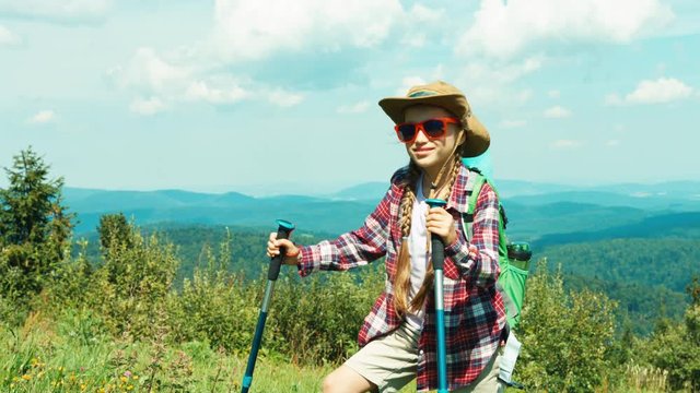  Little hiker girl 7-8 years in sunglasses standing on mountains background smiling at camera and goes up to hill