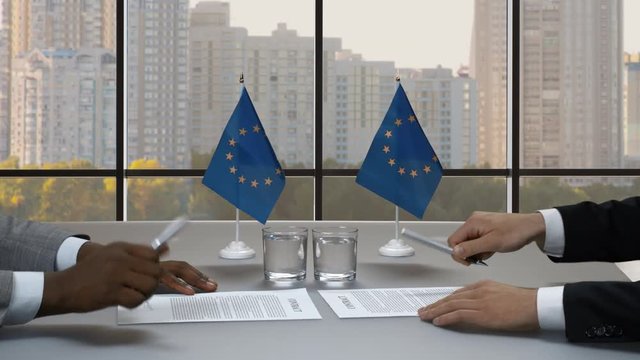 Hands signing papers at desk. Businessmen shake hands near flags. Summit of European Union. Trust and cooperaion.