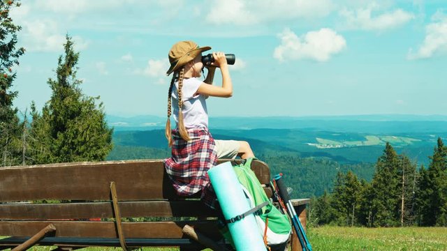 Little hiker girl 7-8 years old using binoculars and sitting on the bench on mountains background and laughing at camera