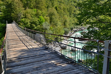 Obraz premium Detail of a wooden foot bridge over a mountain river with blue-green cold water, big stones and freen forest around