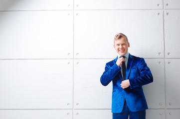 Confident and charisma. Portrait of happy young man in blue suit holding hands on tie looking at camera while standing against white wall.