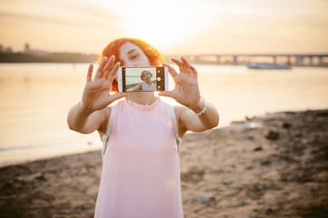 girl in pale pink dress with red hair and backpack walking along river bank, pictures of themselves on their mobile camera phone, against backdrop of boats moored on a warm summer day