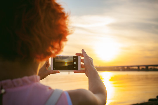 girl in pale pink dress with red hair and backpack walking along river bank, pictures of themselves on their mobile camera phone, against backdrop of boats moored on a warm summer day