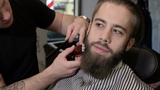 Young bearded man getting beard haircut by hairdresser at barbershop