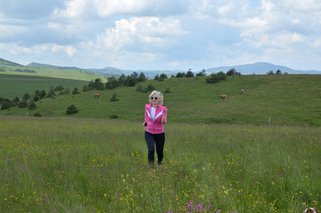 Woman is running on meadow on Zlatibor Mountain in Serbia, with headphones and with hills and cows in background, on a cloudy day