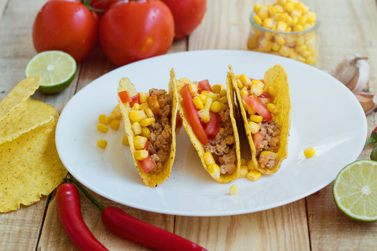 Three crispy tacos with chicken mince, tomatoes and corn served on a white plate together with other ingredients
