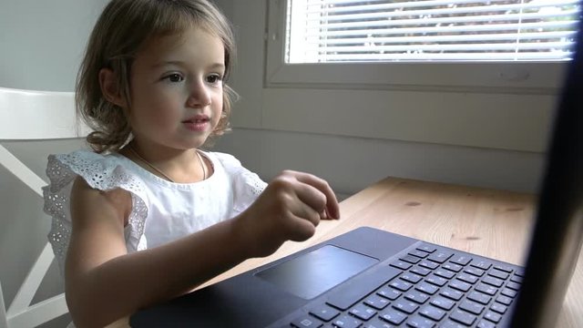 Little girl enthusiastically uses the laptop sitting at the table. dolly shot. Slow motion.
