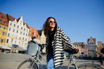 The dark-haired beautiful girl with bicycle on the paved square of the ancient city