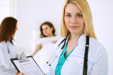 Fototapeta premium Young beautiful female doctor smiling on the background with patient and his physician in hospital. High level and quality medical service concept.