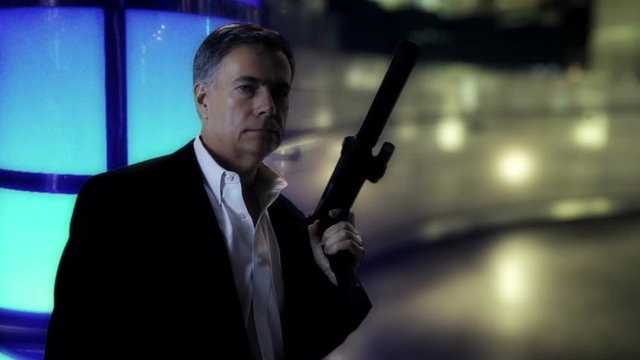 A cagey character standing in a strangely lit room reveals a MP7 firearm he is holding then scans his surroundings.