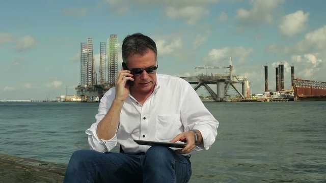 A Man Sitting On A Pier With Drilling Platforms In The Background Using A Cell Phone And Electronic Tablet To Conduct Business.