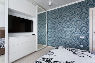 The corner of a bedroom with white commode,tv and a mirror, blue wallpaper
