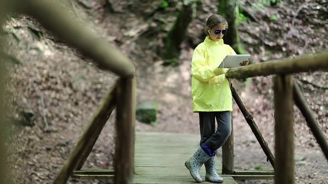 Girl in yellow jacket stands on bridge and uses tablet computer. Pretty girl looks and flips through the photos in his tablet pc. Girl in windcheater with white tablet pc stands on wooden foot bridge