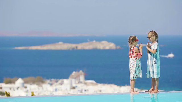 Adorable little girls playing in the edge of outdoor swimming pool with amazing view of old Mykonos town, Europe