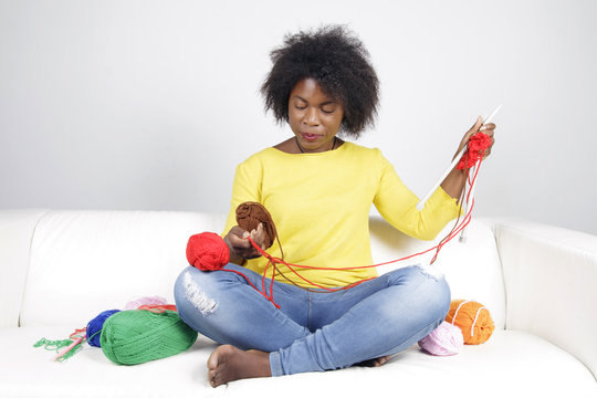 African woman knitting, sitting on a white couch. Surrounded by balls of yarn, she is mixing brown with red colour.