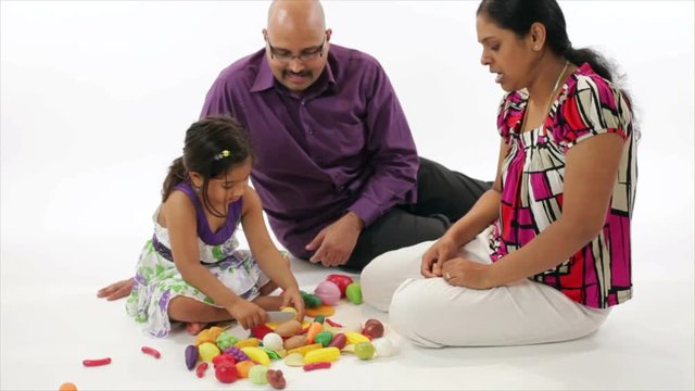 A family of Indian heritage sitting on a white backdrop playing with their cute little daughter.