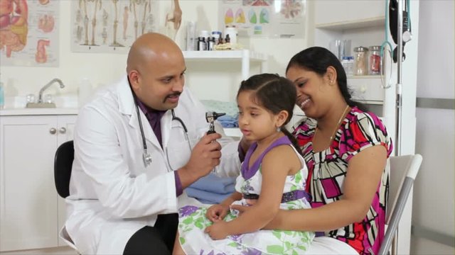An Indian pediatrician or family doctor uses an otoscope to examine the ears of a little girl who is being accompanied by her mother.
