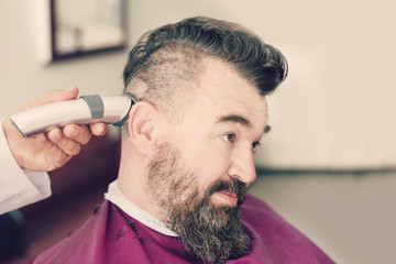 Male barber makes a mohawk hairstyle using clipper of a adult man with beard