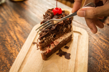 Woman hand use frok to cutting the Slice of chocolate cake with cherries topping on wood table.