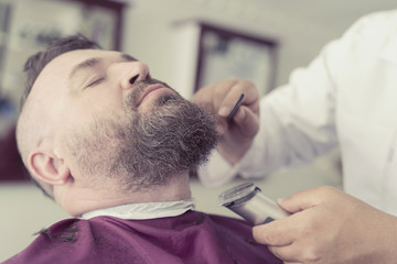 Male barber cuts the beard using clipper of a adult man with a mohawk. Toned