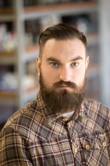 Obraz premium Headshot portrait of serious young man with long beard and mustache. Handsome bearded man posing with hands crossed on his chest, looking at camera, standing in his shop
