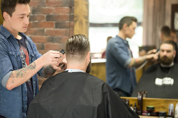 Back view of handsome young bearded man getting trendy haircut in modern barbershop. Cool male hairstylist with tattoo "born barber" serving client. Indoors shot. Incidental People on the background