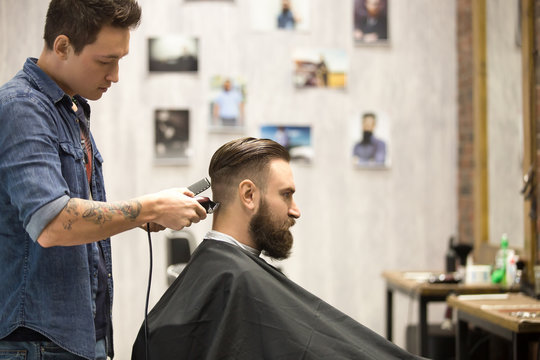 Side view portrait of handsome young bearded caucasian man getting trendy haircut in modern barbershop. Cool men hair stylist with tattoo "born barber" serving client. Indoors shot