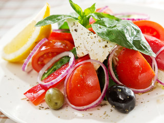 Greek salad with feta cheese slice, tomato, paprika, pepper, onion, cucumber, olives, lemon and basil leaves close up view