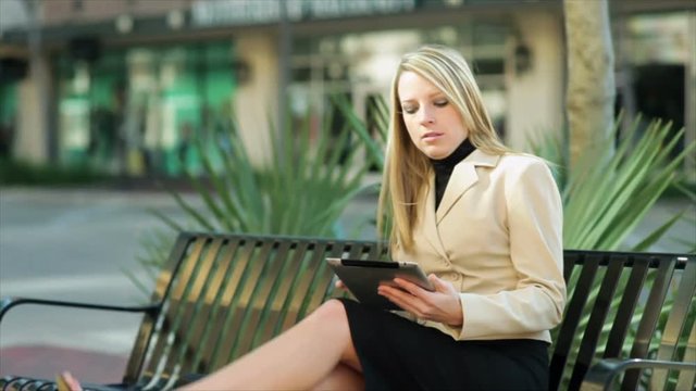 A pretty blond businesswoman walks to and sits on a park bench and uses her electronic tablet.