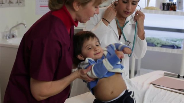 A nurse holds a playful toddler while a pretty Hispanic pediatrician patiently tries to examine him.