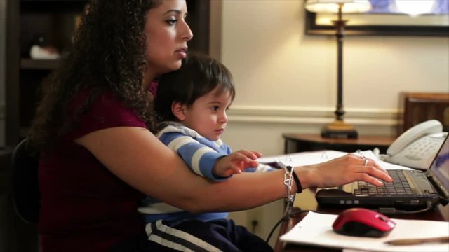 A pretty Hispanic businesswoman in her home office trying to work while holding her young son.