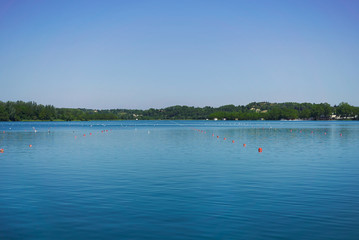 Fototapeta na wymiar Rowing lane marked by orange and white buoys on a peaceful lake surrounded by hills under a clear blue sky