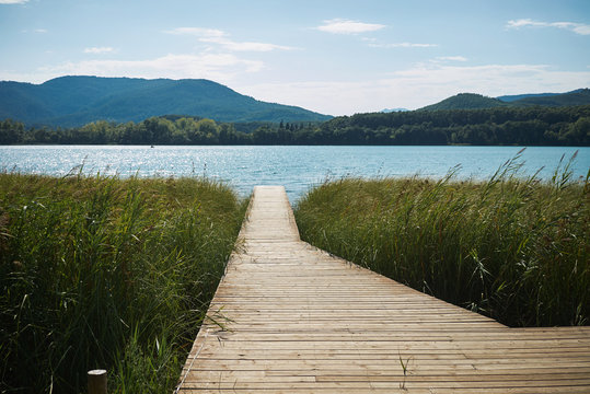 Fototapeta Long wooden pier leads through tall grass to a sparkling blue lake surrounded by wooded hills on a clear summer day