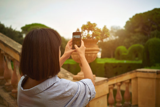 Close up back shot of a dark-haired slim girl taking a photo of park buildings on her smartphone