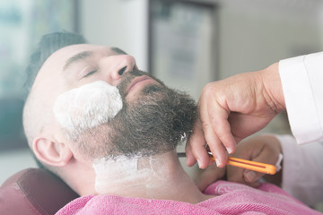 Client with foam on face during beard shaving in barber shop. Toned