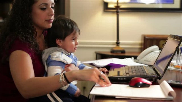 A pretty Hispanic work-at-home mom working while holding her little son.