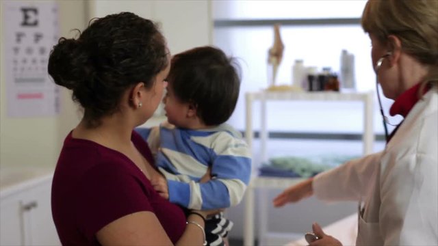 A pretty Hispanic mother holds her cute little toddler as a mature Caucasian pediatrician tries to examine him.