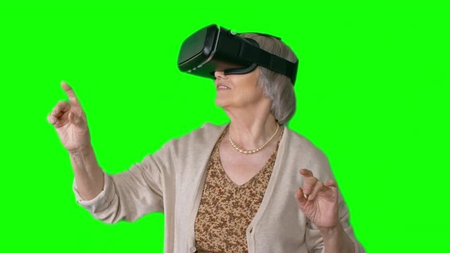 Smiling elderly woman wearing VR goggles standing against green screen background pointing and grabbing something 